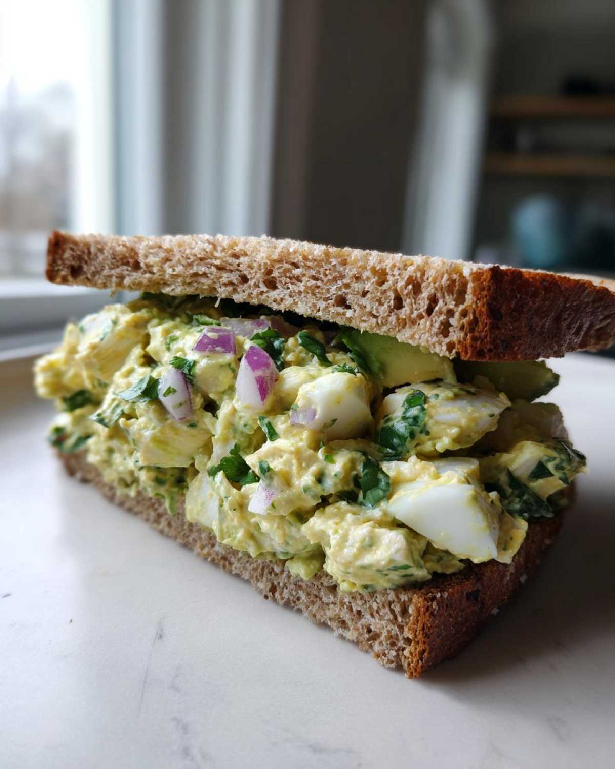 Close-up of a hearty Avocado Egg Salad Sandwich made with whole wheat bread, visible chunks of egg, and red onion.