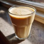 A tall glass filled with a thick, frothy Banana Peanut Butter Smoothie sitting on a sunlit countertop.