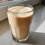 A tall glass filled with a creamy Banana Peanut Butter Smoothie sitting on a bright countertop near a window.