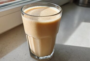 A tall glass filled with a creamy Banana Peanut Butter Smoothie sitting on a bright countertop near a window.