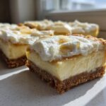 A close-up, sunlit view of a thick slice of Banana Pudding Cheesecake Bars showing the graham cracker crust, creamy filling, and whipped topping.
