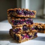 A stack of four moist Blueberry Baked Oatmeal Squares showing the thick, purple blueberry filling between the oat layers.