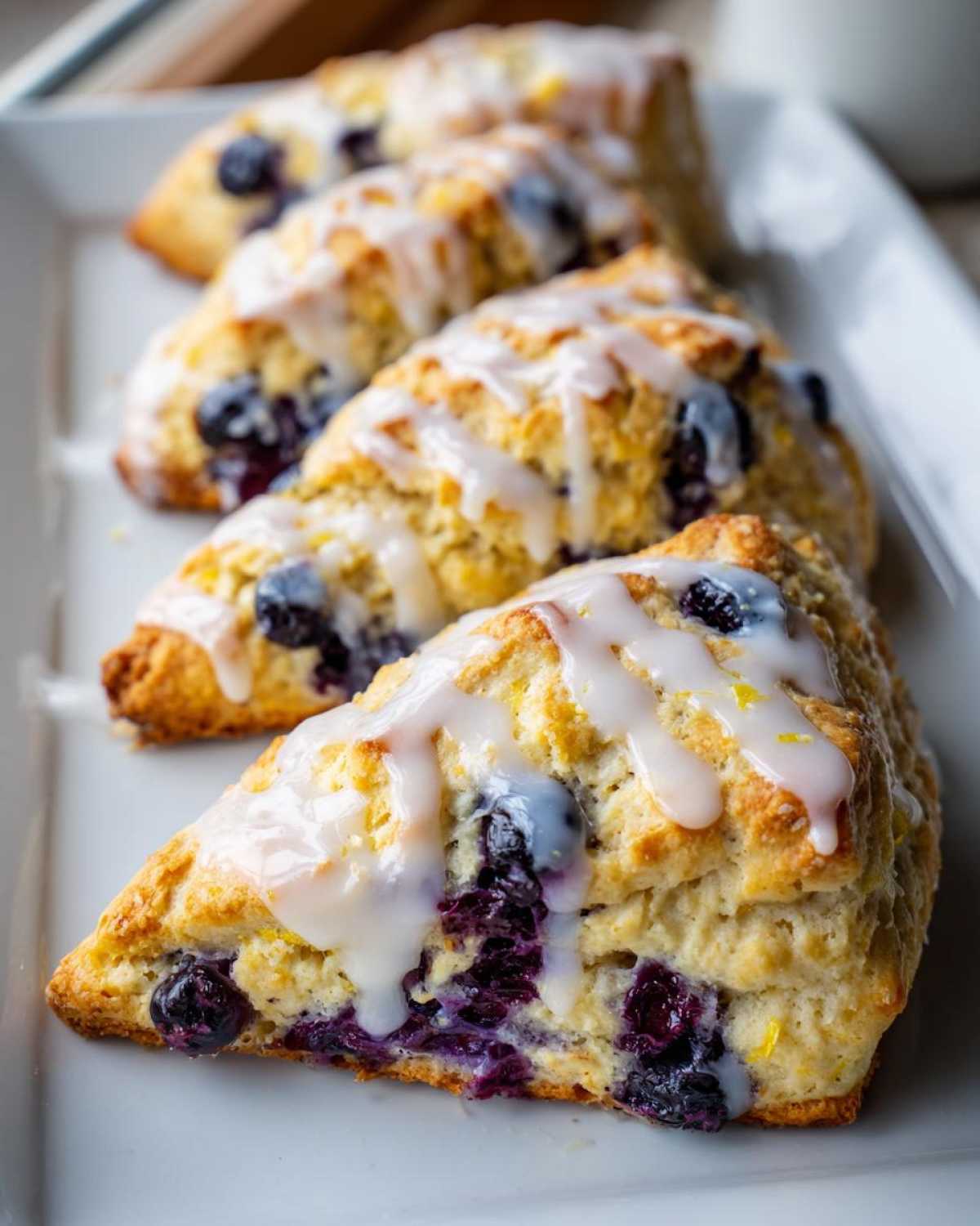 Close-up of several triangular Blueberry Lemon Scones drizzled with white lemon glaze, showing bursts of purple blueberries.