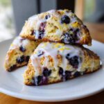Three delicious Blueberry Lemon Scones stacked on a white plate, topped with white glaze and lemon zest.