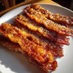 Close-up of crispy, caramelized Brown Sugar Glazed Bacon strips arranged on a white plate, glistening in the sunlight.