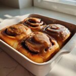 Four freshly baked Gluten-Free Cinnamon Rolls topped with thick caramel icing in a white baking dish.