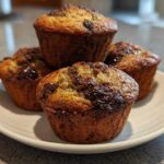 A stack of four freshly baked, moist Chocolate Chip Banana Muffins with melted chocolate topping on a white plate.