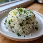 A mound of fluffy white Cilantro Lime Rice topped with chopped cilantro and yellow lime zest on a light plate.