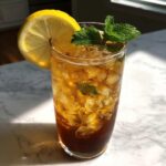 Tall glass filled with ice and dark amber Cold Brew Lemonade, garnished with a lemon slice and fresh mint.