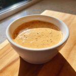 A small white bowl filled with creamy, orange-hued Copycat Raising Cane’s Sauce speckled with black pepper.