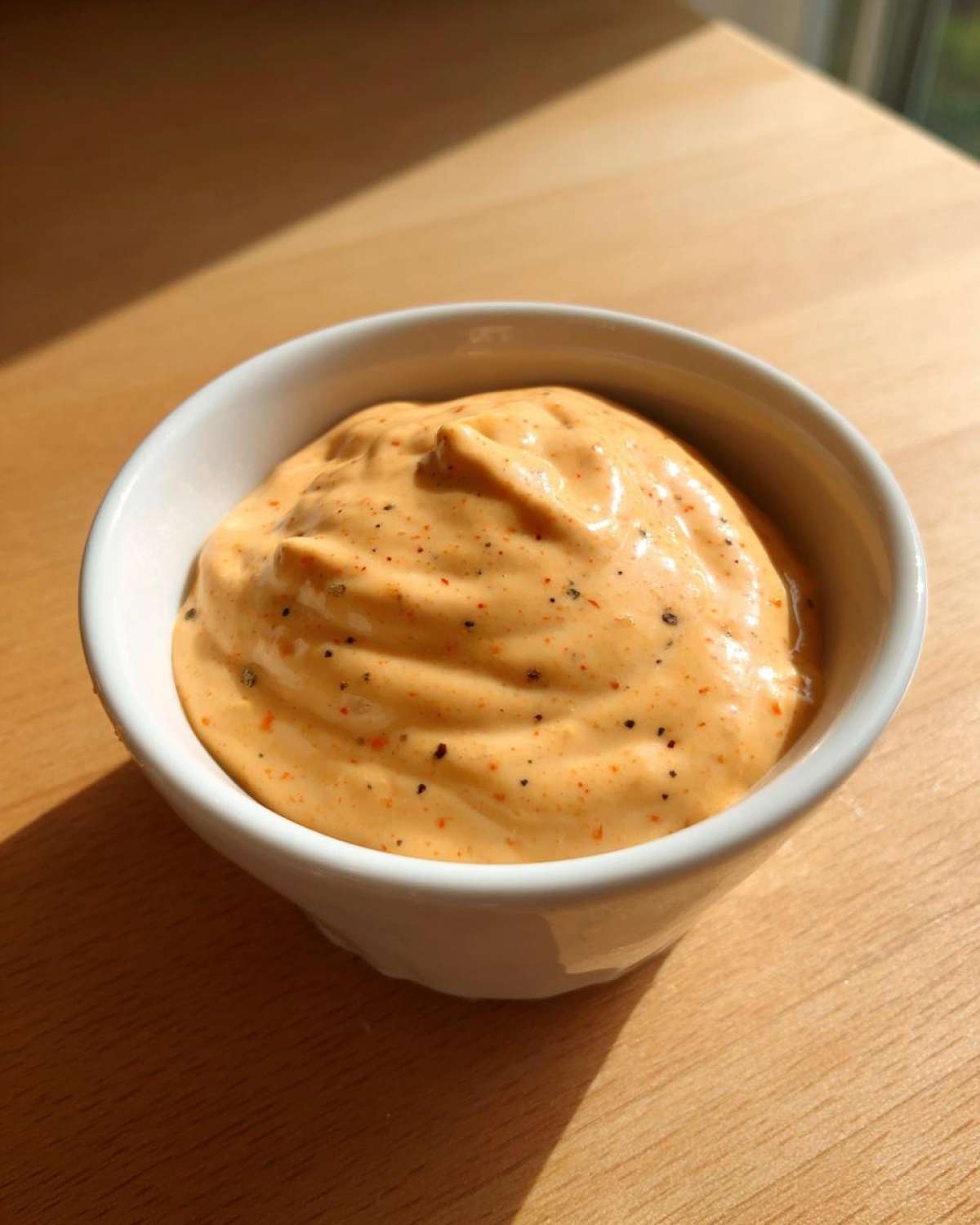 A close-up of thick, orange Copycat Raising Cane’s Sauce speckled with spices in a small white dipping bowl.