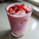 A tall glass filled with a swirling pink and white Copycat Starbucks Pink Drink, topped with ice and dried strawberry pieces.