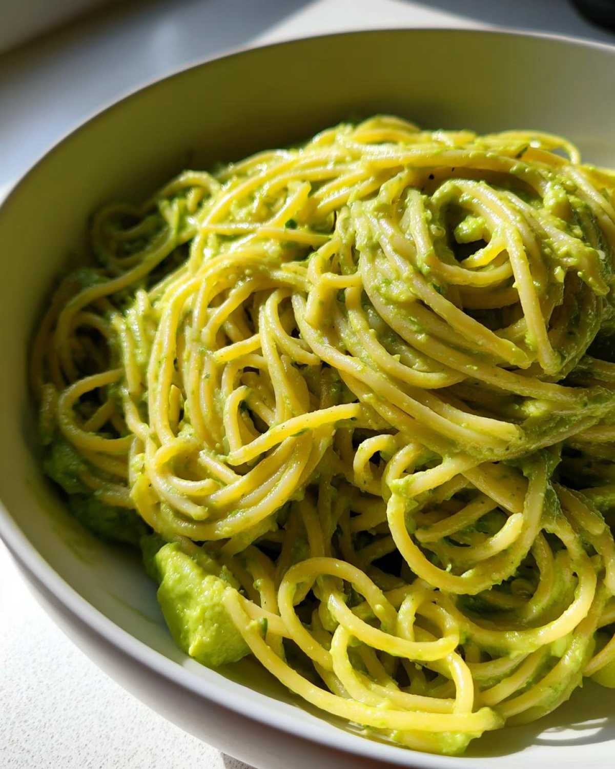 A close-up shot of spaghetti coated in vibrant green, creamy avocado pasta sauce served in a light green bowl.