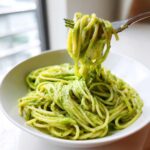 A fork lifting a twirl of spaghetti coated in vibrant green Creamy Avocado Pasta sauce from a white bowl.
