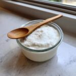 A glass jar filled with a thick, white creamy base ingredient for Copycat Olive Garden Salad Dressing, topped with a small wooden spoon.