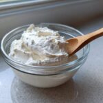 A glass bowl filled with thick, creamy white dressing, likely the base for Copycat Olive Garden Salad Dressing, with a wooden spoon resting in it.