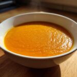 Close-up of a white bowl filled with hot, vibrant orange Creamy Coconut Lentil Soup on a wooden surface.