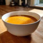 A steaming white bowl filled with bright orange Creamy Coconut Lentil Soup, sitting on a wooden countertop in sunlight.