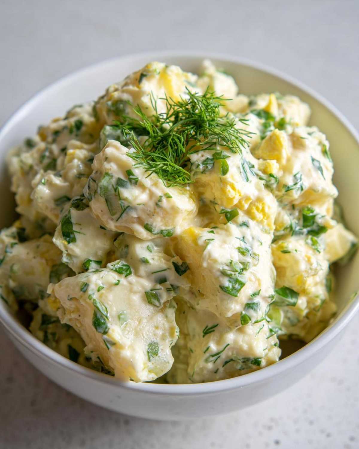 A close-up of a white bowl filled with chunky Creamy Dill Potato Salad, garnished with fresh dill.