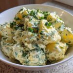 A close-up of chunks of boiled potato coated in a creamy dressing, heavily mixed with fresh dill and chives, served in a white bowl. This is the Creamy Dill Potato Salad.