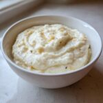 A white bowl filled with smooth, creamy garlic mashed cauliflower, garnished with browned garlic bits.