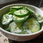 Close-up of a white bowl filled with slices of Creamy German Cucumber Salad tossed in a white dressing and topped with fresh dill.