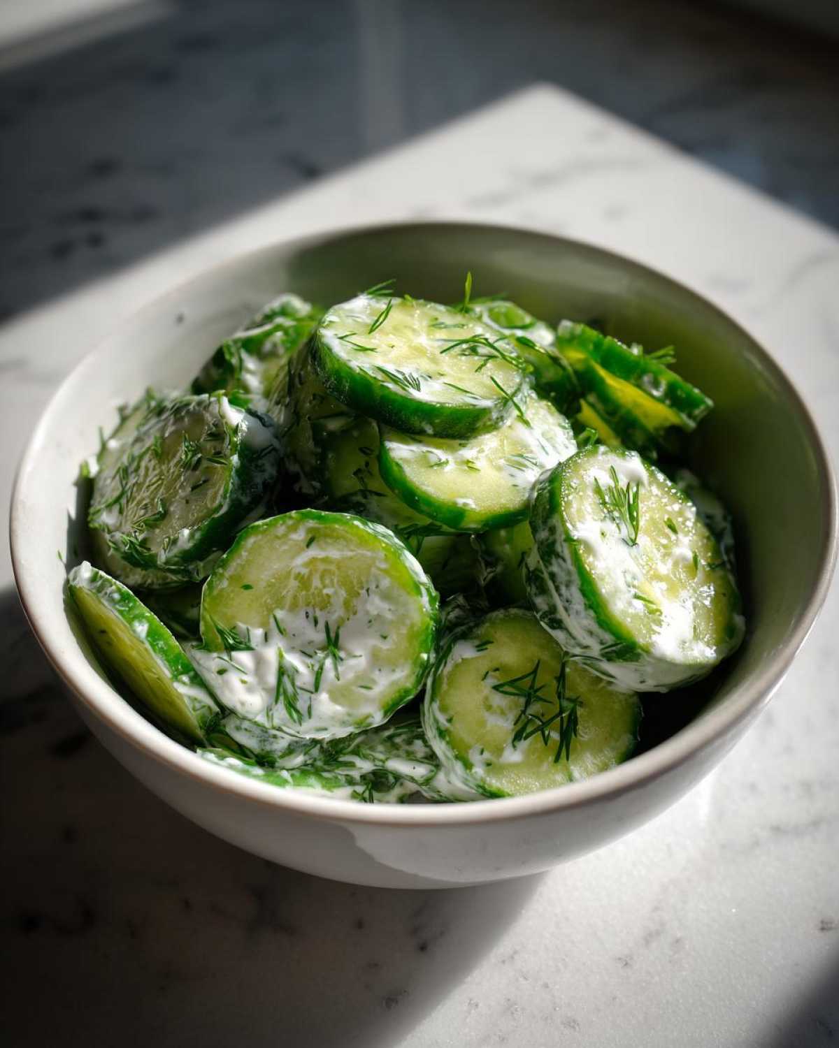Close-up of sliced cucumbers coated in a creamy dressing and fresh dill, served in a bowl of Creamy German Cucumber Salad.