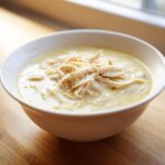 A close-up of a white bowl filled with rich Creamy Lemon Chicken Orzo Soup topped with shredded chicken.