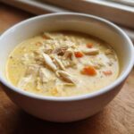 Close-up of a white bowl filled with Creamy Lemon Chicken Orzo Soup, featuring shredded chicken and visible carrot pieces.