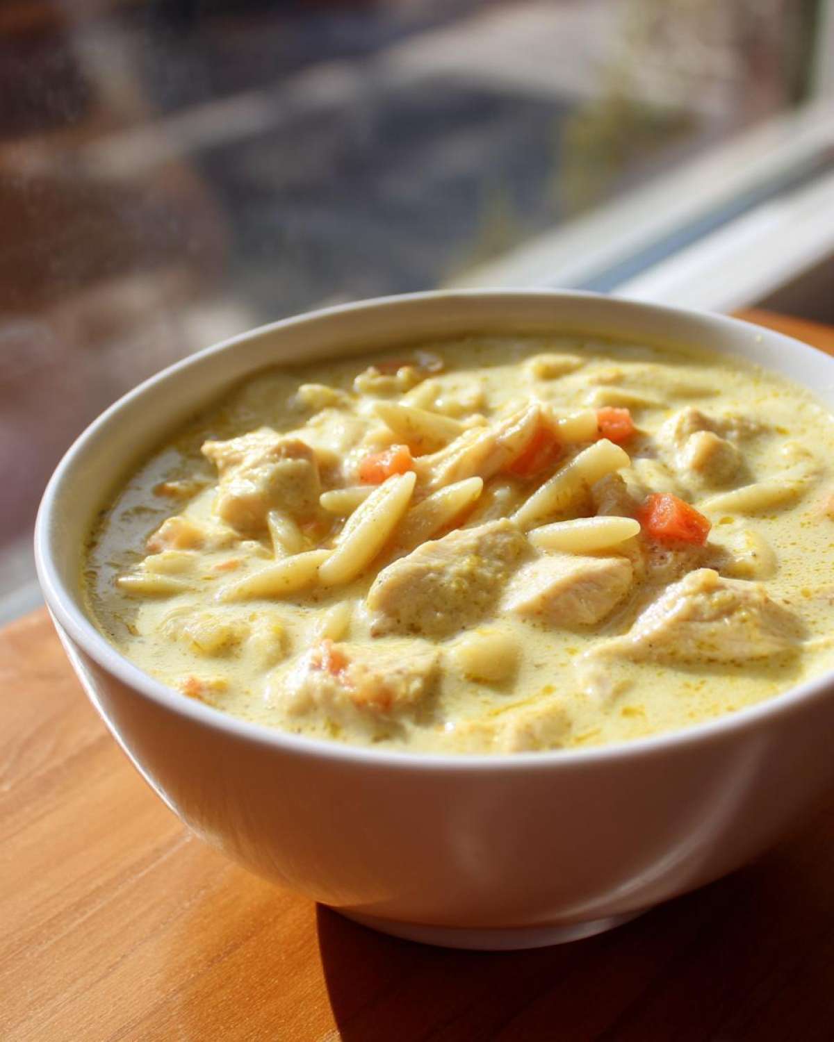 Close-up of a white bowl filled with steaming Creamy Lemon Chicken Orzo Soup, showing chicken chunks, orzo pasta, and carrots.