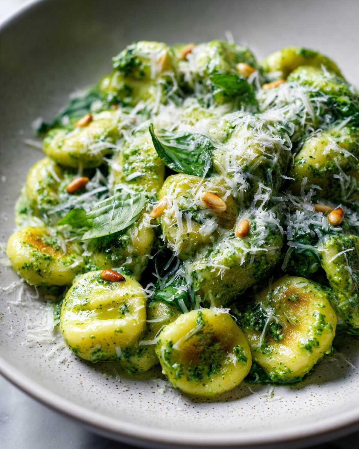 A close-up bowl of tender, pan-seared Creamy Pesto Gnocchi topped with shredded Parmesan cheese and pine nuts.