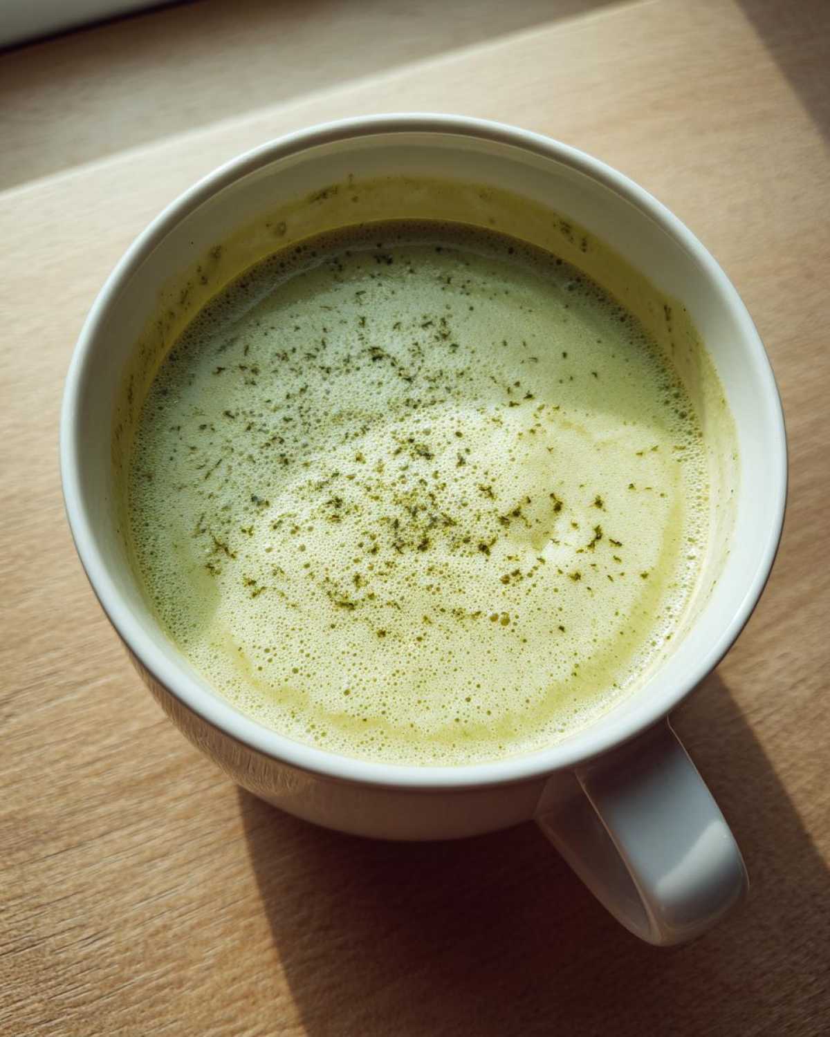 Close-up of a light green, foamy beverage, possibly Creamy Potato Leek Soup, in a white mug on a wooden surface.