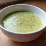Close-up of a white mug filled with light green, creamy potato leek soup, garnished with herbs.