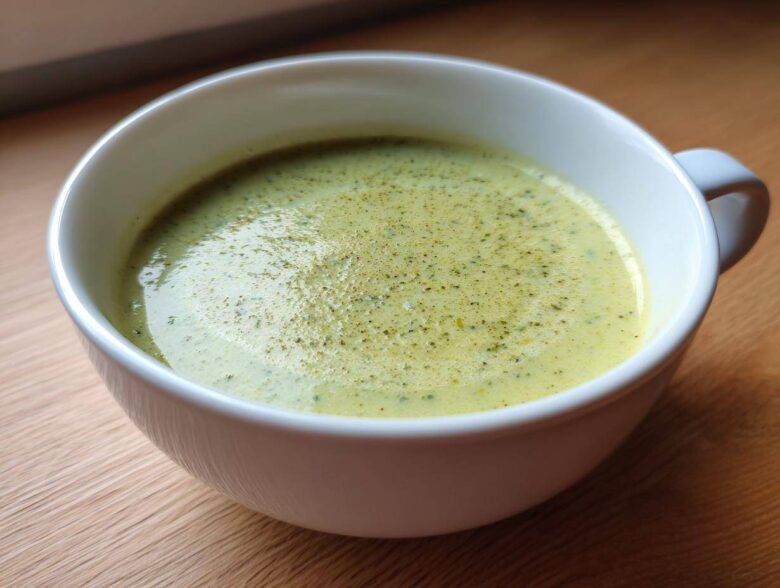 Close-up of a white mug filled with light green, creamy potato leek soup, garnished with herbs.