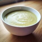 Close-up of a white bowl filled with hot, creamy potato leek soup, lightly seasoned with pepper.
