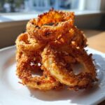 A close-up stack of golden brown, crispy Air Fryer Onion Rings resting on a white plate, catching bright sunlight.