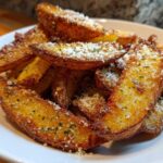 A close-up of golden brown Crispy Baked Garlic Parmesan Potato Wedges piled high on a white plate, dusted with cheese.