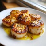 Close-up of six perfectly seared Garlic Butter Scallops with Lemon sauce pooling on a white plate.