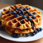 A close-up of a stack of golden-brown Gluten-Free Blueberry Waffles topped generously with fresh blueberries.