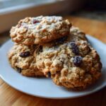 A stack of four freshly baked Gluten-Free Oatmeal Raisin Cookies dusted with powdered sugar on a white plate.