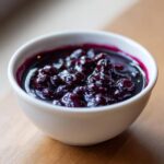 Close-up of rich, dark purple Homemade Blueberry Syrup with whole berries in a small white bowl.