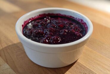 Close-up of thick, deep purple Homemade Blueberry Syrup with whole berries in a small white bowl.