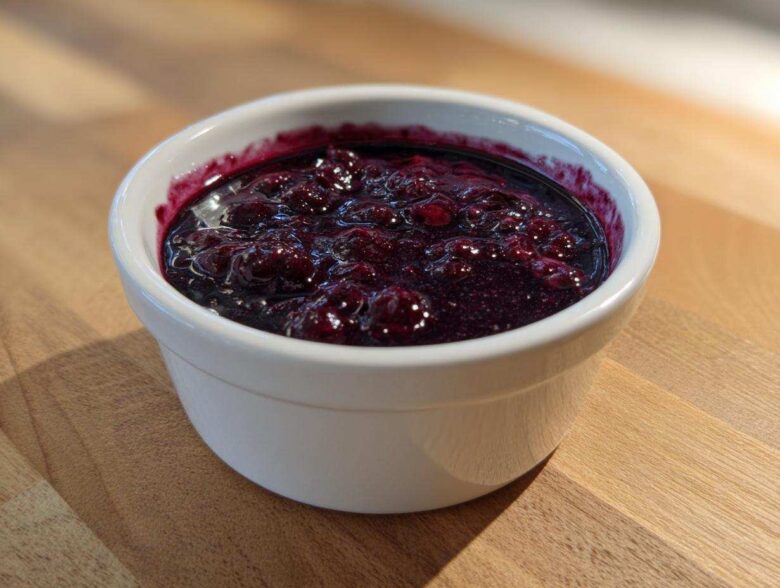 Close-up of thick, deep purple Homemade Blueberry Syrup with whole berries in a small white bowl.