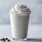 A tall glass of Homemade Vanilla Bean Frappuccino topped with whipped cream and coffee beans nearby.