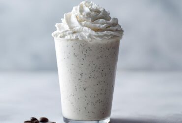 A tall glass of Homemade Vanilla Bean Frappuccino topped with whipped cream and coffee beans nearby.