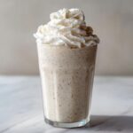 A tall glass filled with a speckled Homemade Vanilla Bean Frappuccino topped generously with whipped cream.