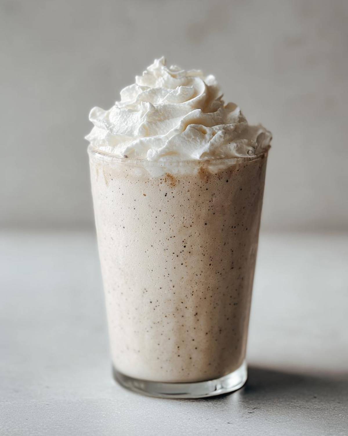 A tall glass filled with a creamy Homemade Vanilla Bean Frappuccino, topped generously with whipped cream.