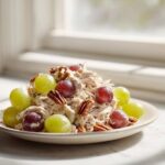 A serving of Honey Mustard Chicken Salad with Grapes and pecans on a light plate near a window.