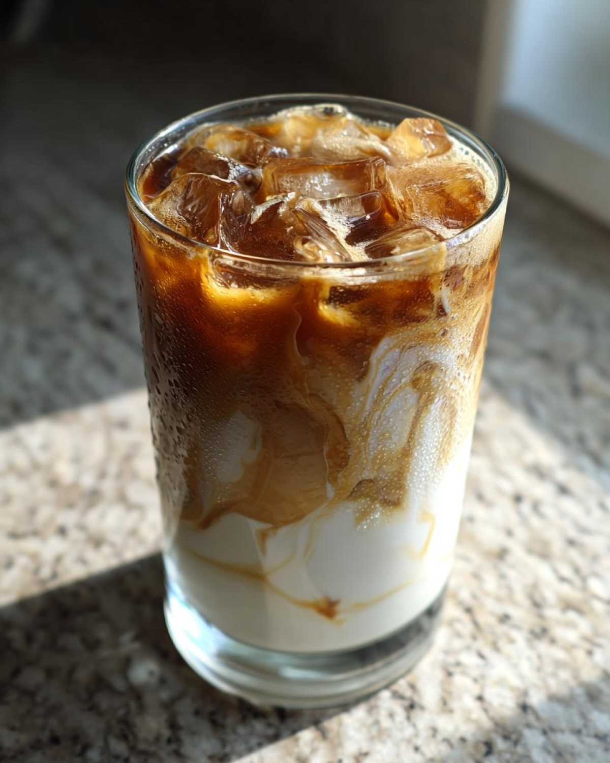 Close-up of a tall glass containing an Iced Honey Almond Milk Latte with swirling coffee and milk layers over ice.
