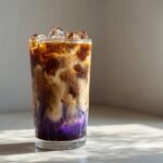 Close-up of a tall glass containing a layered Iced Lavender Latte with purple syrup, coffee, and cream swirling over ice.